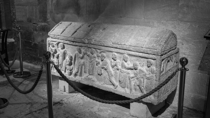 Black and white photo of an intricately carved ancient stone sarcophagus indoors.